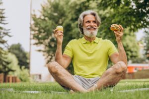 Healthy older man sitting on grass holding an apple and a burger, symbolizing balanced diet choices